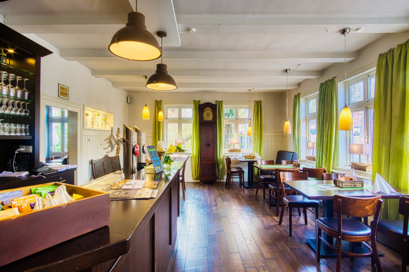 Heller Gastraum mit dunklem Holzboden, Theke, Tischen und grünen Vorhängen an den Sprossenfenstern.