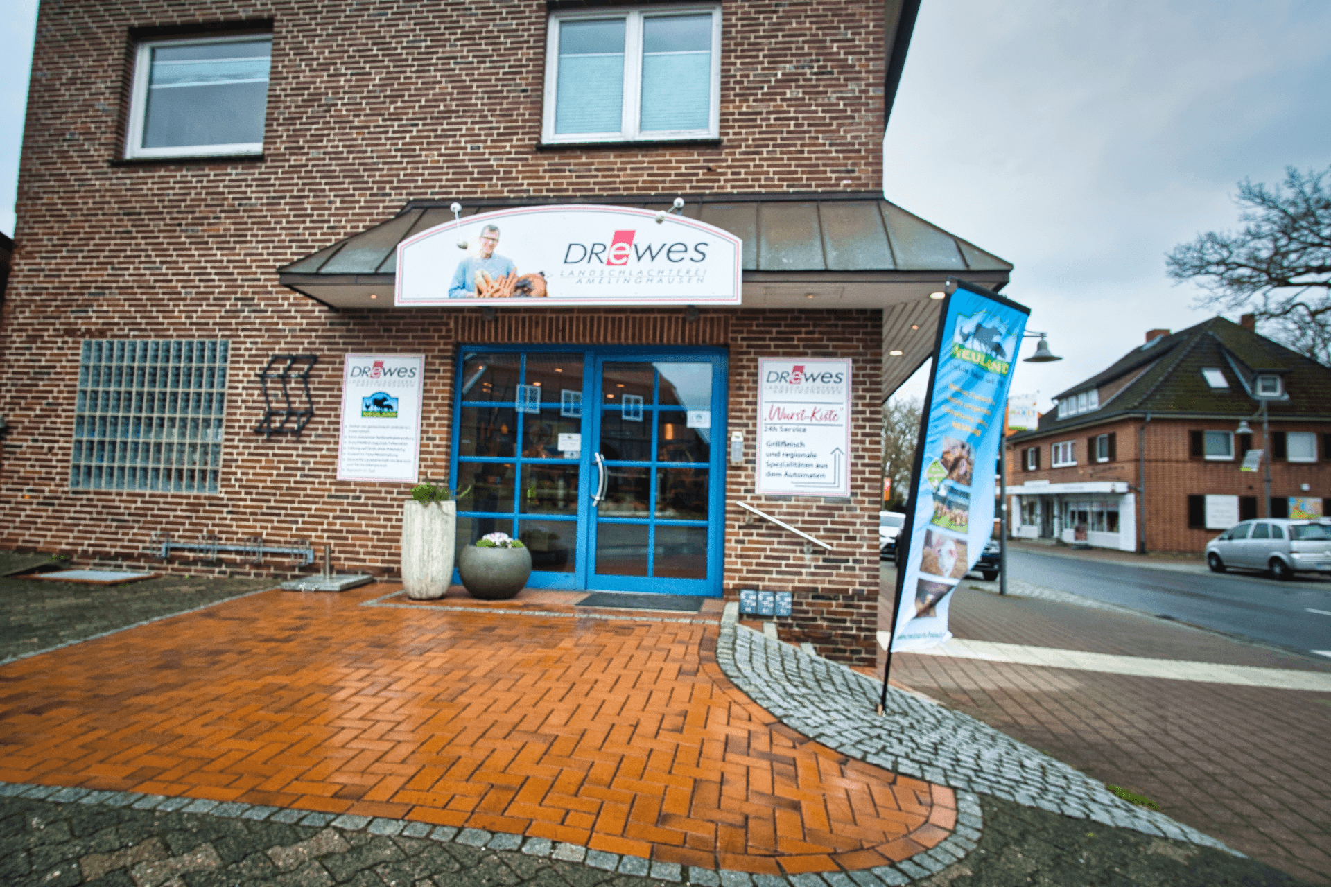 Backsteinfassade der Landschlachterei Drewes mit blauer Eingangstür und Werbeschildern.