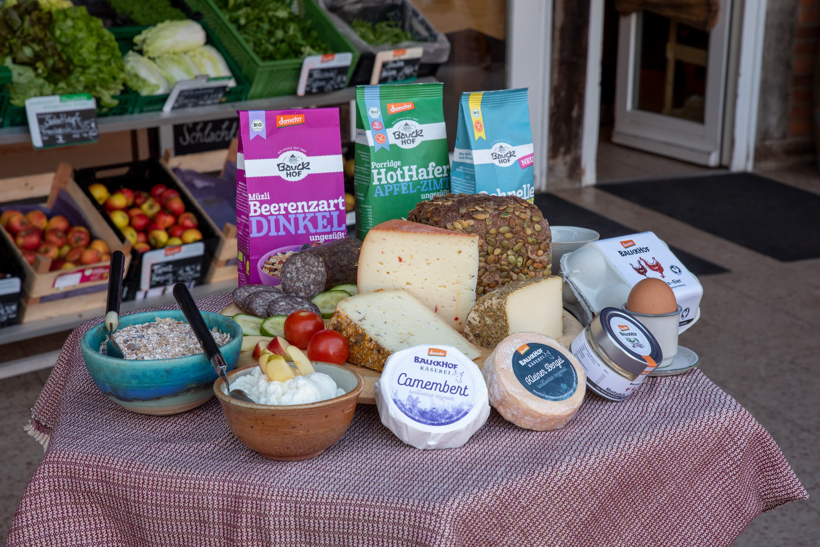 Das Bild zeigt eine liebevoll arrangierte Auswahl an biologischen Backwaren, verschiedenen Käsesorten und Müsli-Schalen, die appetitlich auf einem Tisch drapiert sind. Im Hintergrund sind frisches Bio-Gemüse und Obst zu sehen, die die Vielfalt und Frische des Sortiments unterstreichen.