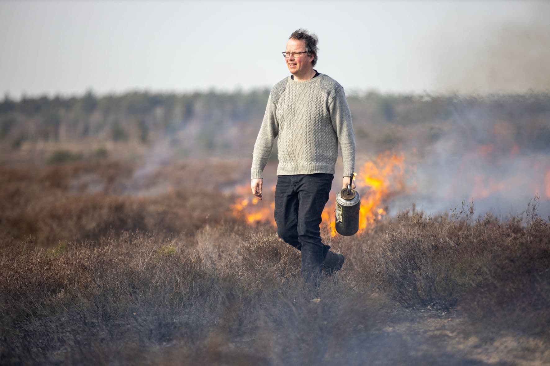 ein mann läuft vor einer brennenden heidefläche, im hintergrund sind lodernde flammen auf dem boden zu sehen