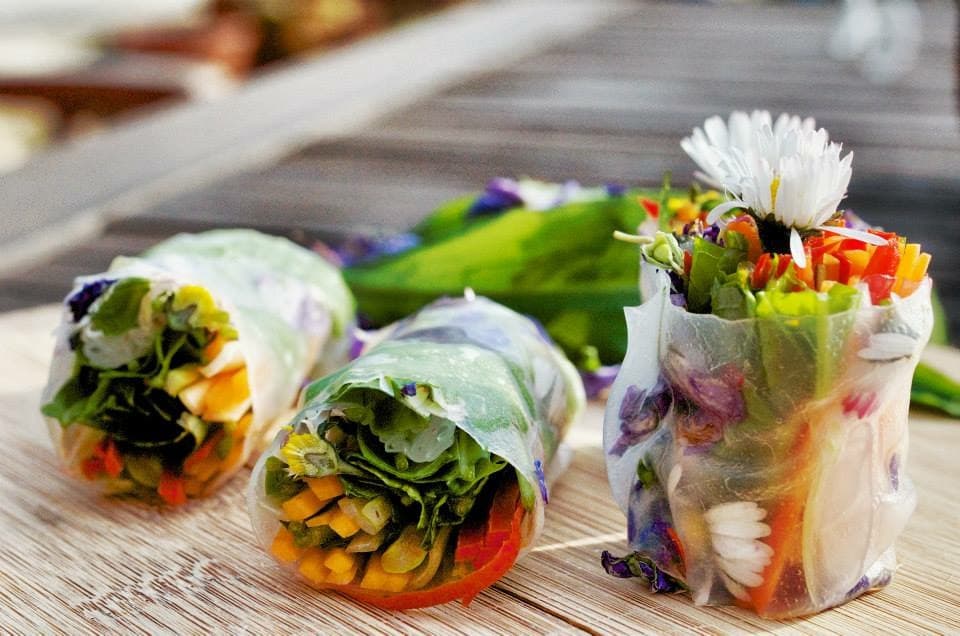 Das Bild zeigt drei frische Frühlingsrollen mit buntem Gemüse und essbaren Blüten auf einem Holztisch.