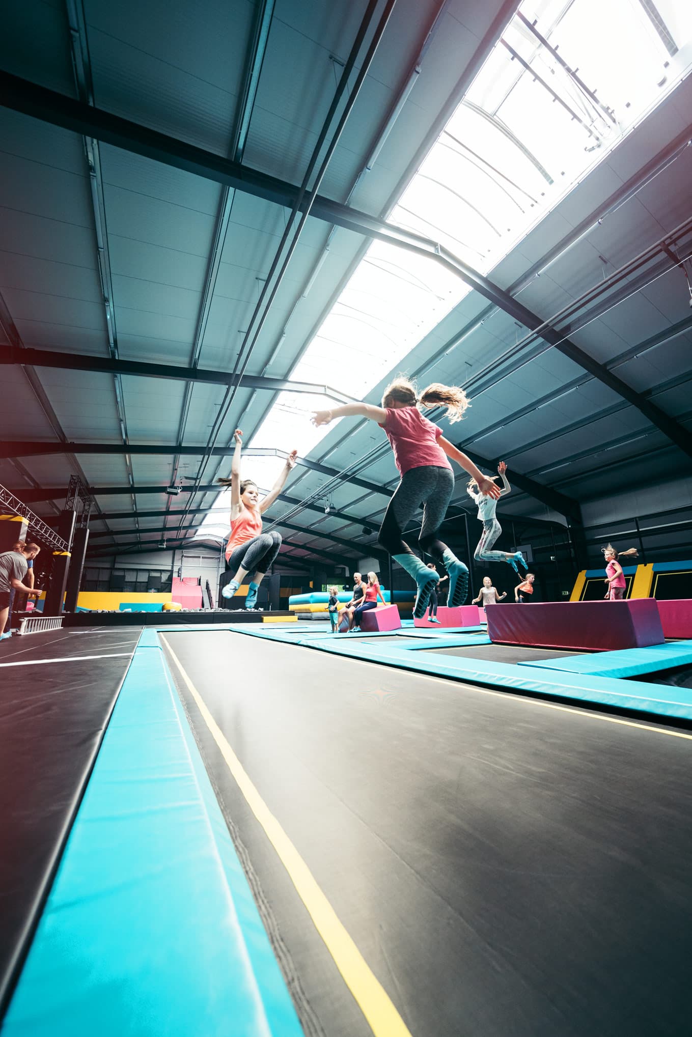 Mehrere Personen springen auf parallel angeordneten Trampolinen in einer hellen Halle.Several people jumping on trampolines arranged in parallel in a bright hall.Flere mennesker hopper på trampoliner, der er arrangeret parallelt i en stærkt oplyst hal.Verschillende mensen springen op trampolines die parallel zijn opgesteld in een helder verlichte hal.