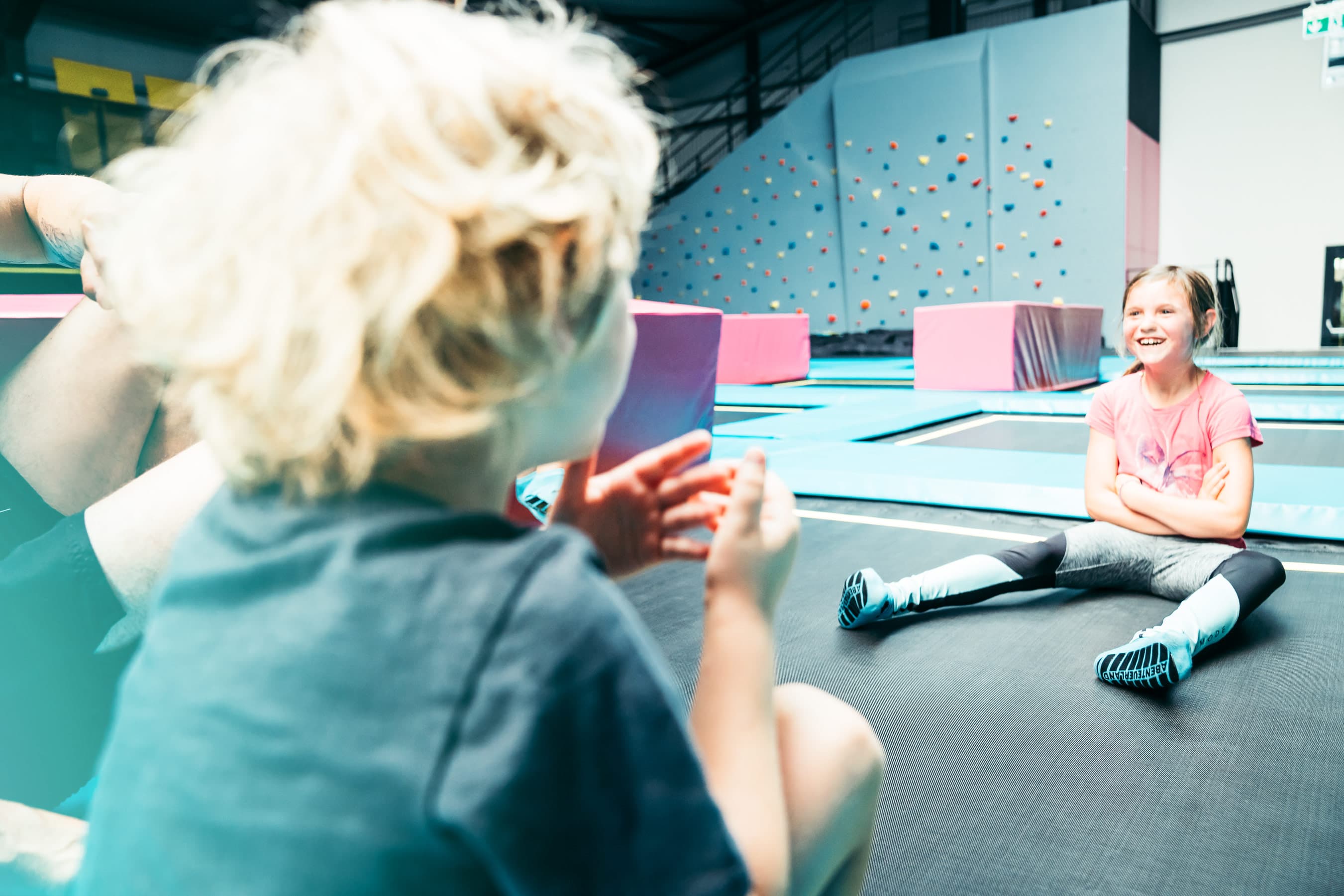 Ein blondes Kind im Fokus sitzt auf einem Trampolin, während ein lächelndes Mädchen im Hintergrund auf dem Trampolin entspannt. Im Raum sind bunte Plattformen und eine Kletterwand zu sehen.A blonde child in focus sits on a trampoline, while a smiling girl relaxes on the trampoline in the background. Colorful platforms and a climbing wall can be seen in the room.Et blondt barn i fokus sidder på en trampolin, mens en smilende pige slapper af på trampolinen i baggrunden. I rummet ses farverige platforme og en klatrevæg.Een blond kind in focus zit op een trampoline, terwijl een lachend meisje ontspant op de trampoline in de achtergrond. In de kamer zijn kleurrijke platforms en een klimwand te zien.