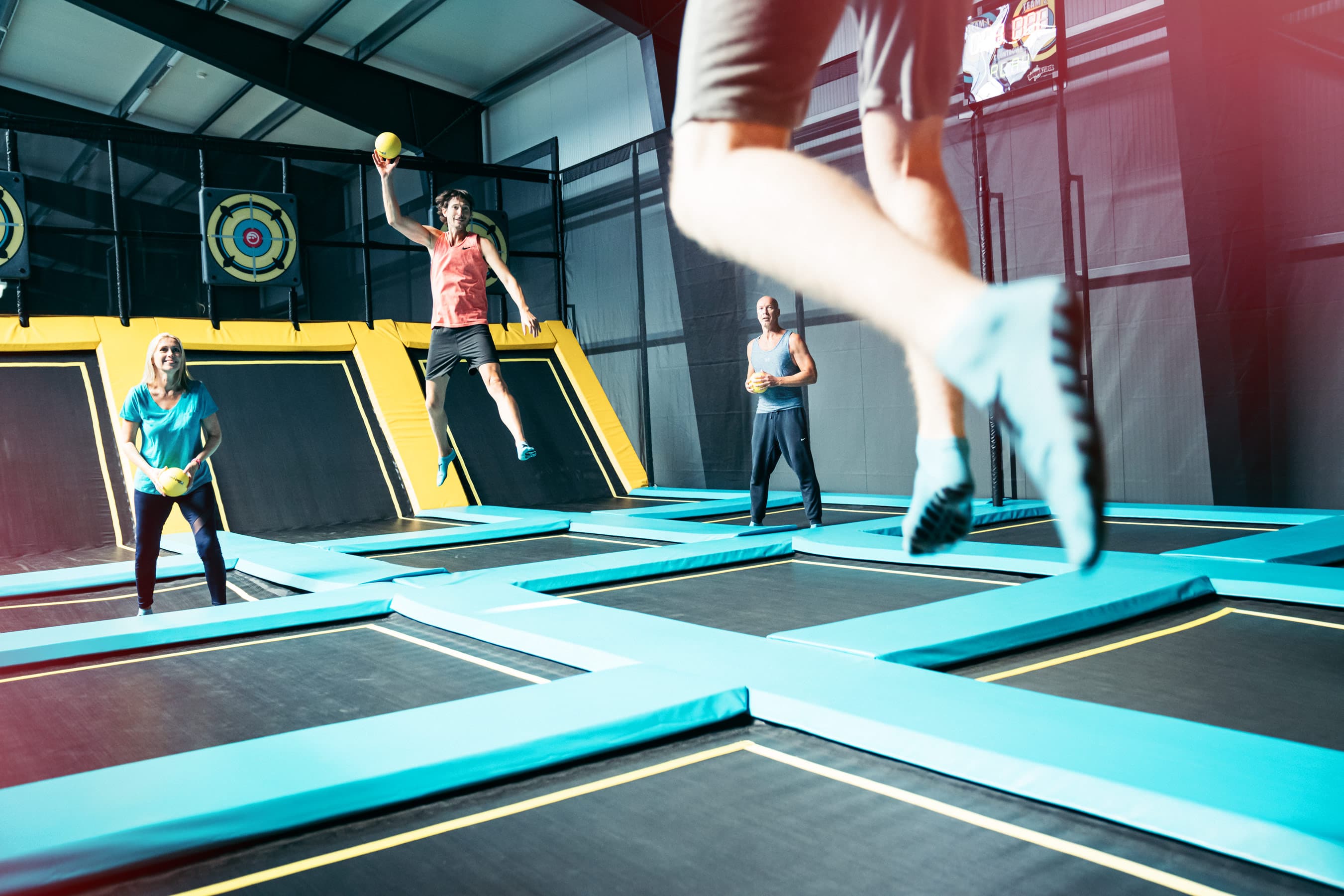 Eine Gruppe von Menschen hat Spaß auf einem Trampolinpark mit bunten Sprungflächen und Zielscheiben. Eine Person springt mit ausgestrecktem Arm und Ball in der Hand.A group of people are having fun on a trampoline park with colorful jumping surfaces and targets. One person is jumping with an outstretched arm and a ball in his hand.En gruppe mennesker har det sjovt i en trampolinpark med farverige springflader og mål. En person hopper med en udstrakt arm og en bold i hånden.Een groep mensen vermaakt zich op een trampolinepark met kleurrijke springoppervlakken en doelen. Eén persoon springt met een uitgestrekte arm en een bal in zijn hand.