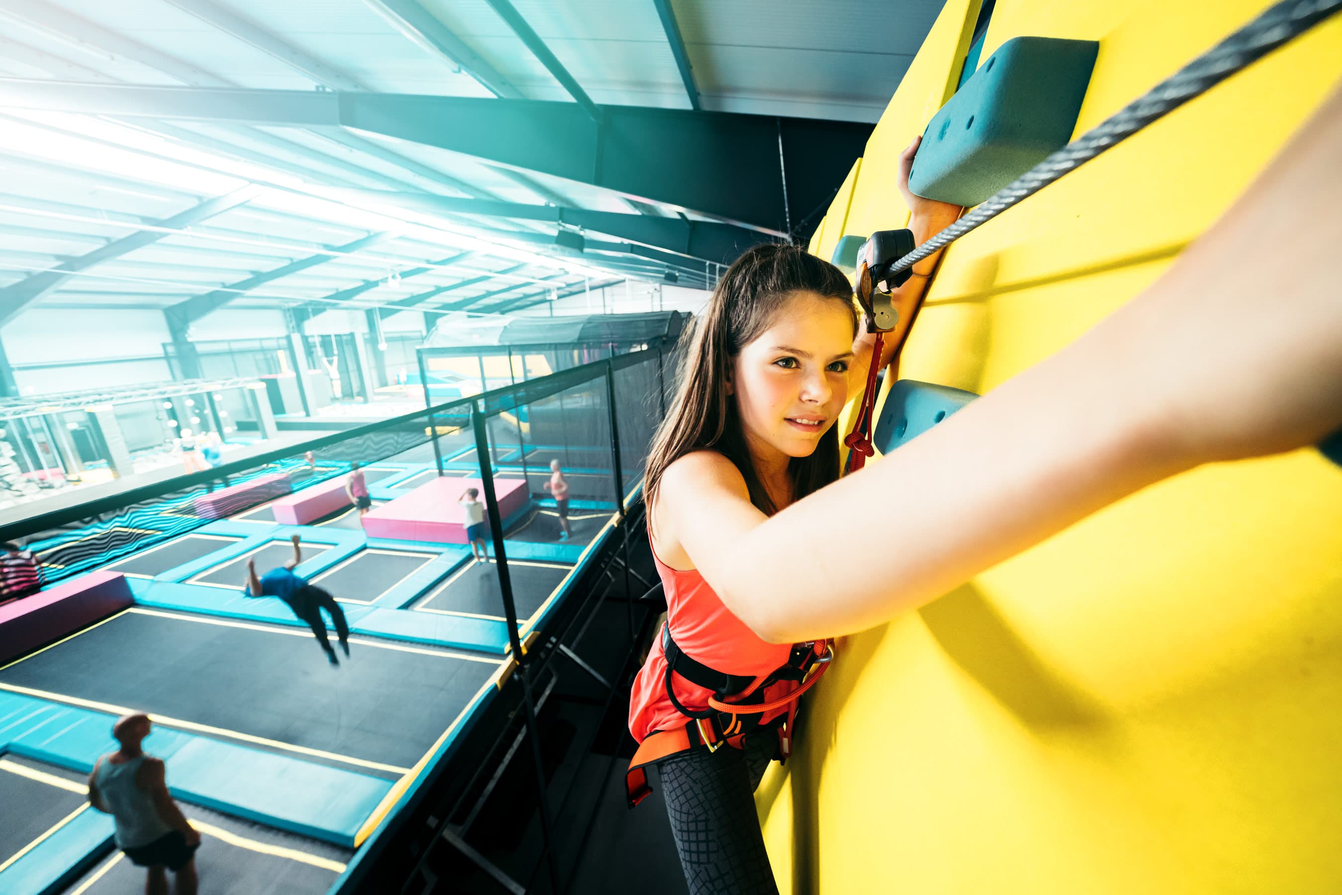 Eine junge Frau erklimmt konzentriert eine gelbe Kletterwand, umgeben von Trampolin-Arealen.A young woman scales a yellow climbing wall surrounded by trampoline areas.En ung kvinde bestiger en gul klatrevæg omgivet af trampolinområder.Een jonge vrouw beklimt een gele klimmuur omringd door trampolines.