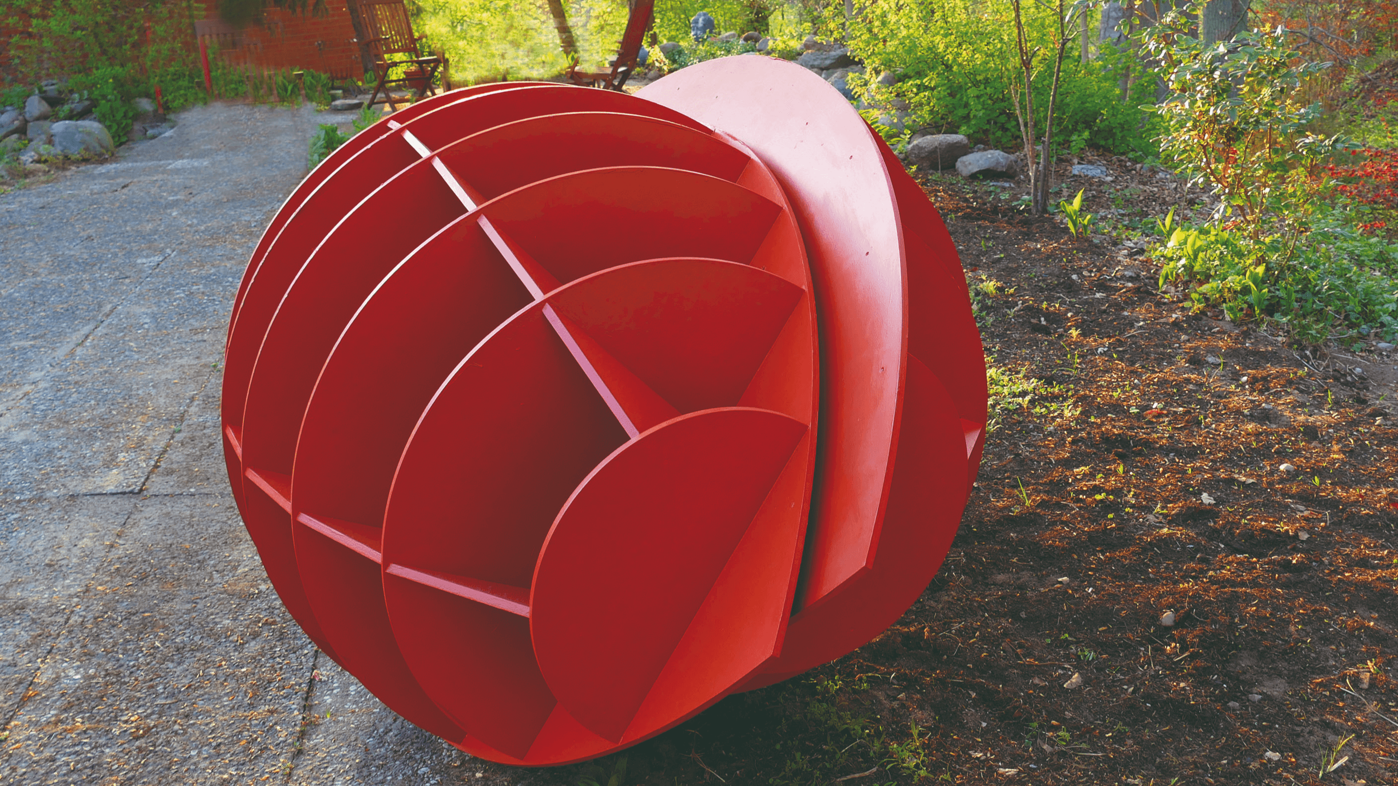 Das Bild zeigt eine kunstvolle, rot bemalte Skulptur im Freien, die kugelförmig gestaltet ist und aus mehreren Segmenten besteht. Sie liegt auf einem gepflasterten Weg und wird von grüner Vegetation umrahmt.