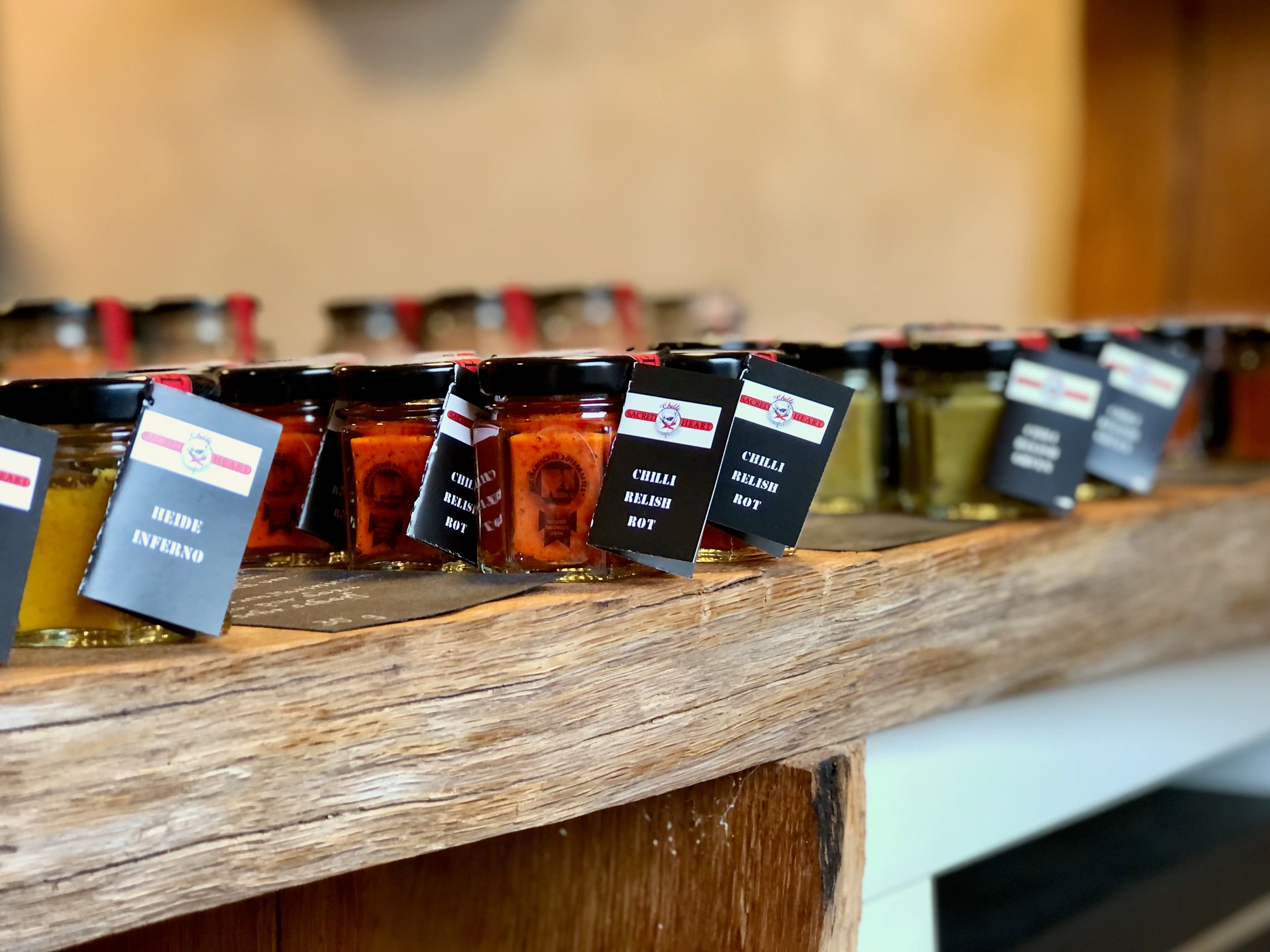 Das Bild zeigt einen Tisch aus Eichenholz im Hofladen. Darauf kleine Gläser mit Chilipasten in unterschiedlichen Geschmacksrichtungen aus eigener Herstellung in der Lüneburger Heide Chilli Manufaktur Susanne Menke Lüneburger Heide