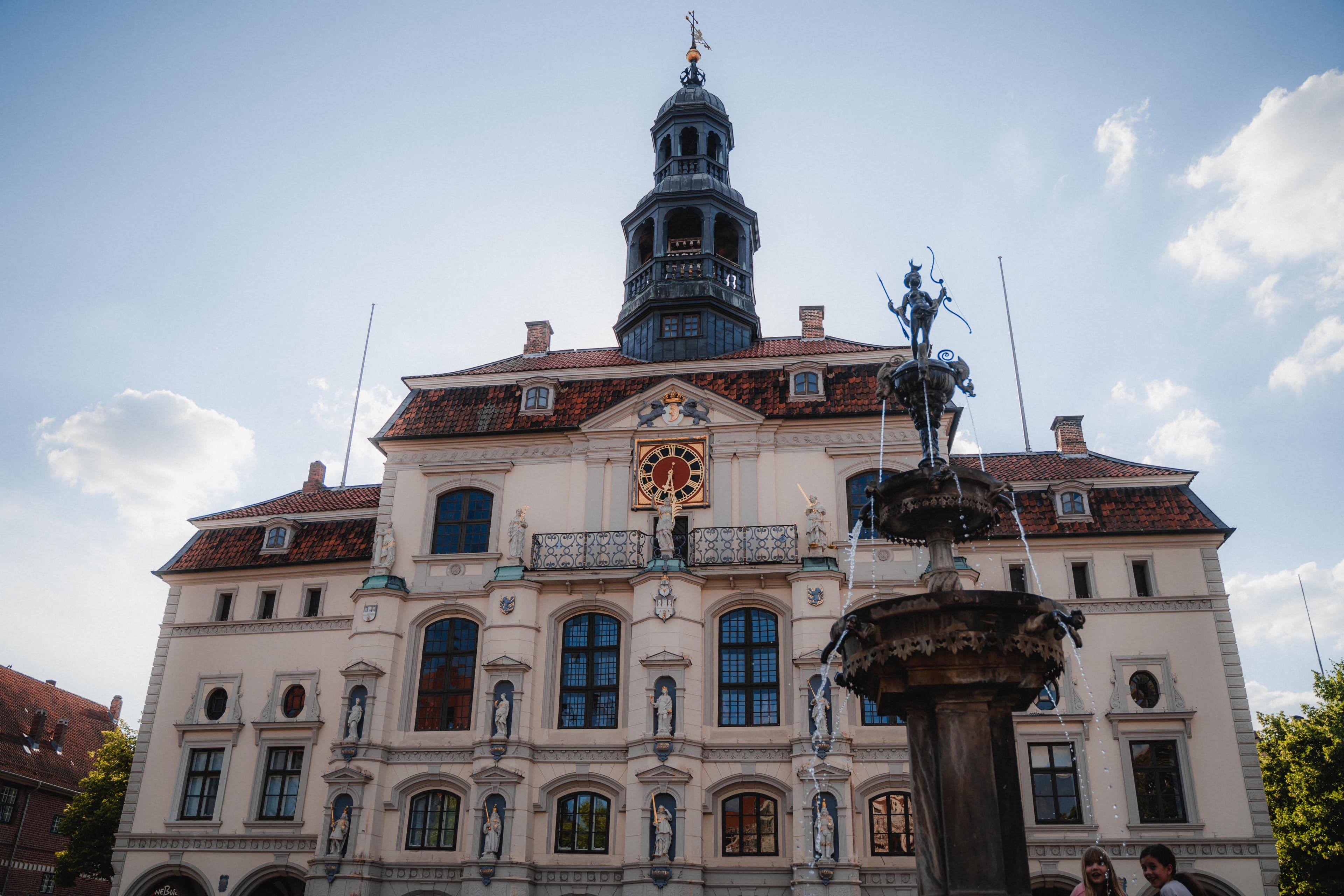 Rathaus Lüneburg mit heller Fassade, Statuen, Uhr und dem Lunabrunnen im Vordergrund.