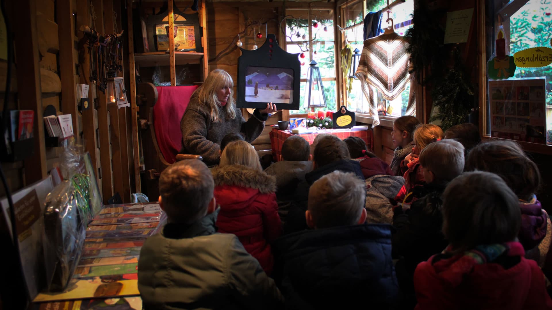 Eine Frau liest einer Gruppe von Kindern in einer hölzernen Hütte auf dem Weihnachtsmarkt vor.A woman reads to a group of children in a wooden hut at the Christmas market.En kvinde læser op for en gruppe børn i en træhytte på julemarkedet.Een vrouw leest voor aan een groep kinderen in een houten hut op de kerstmarkt.