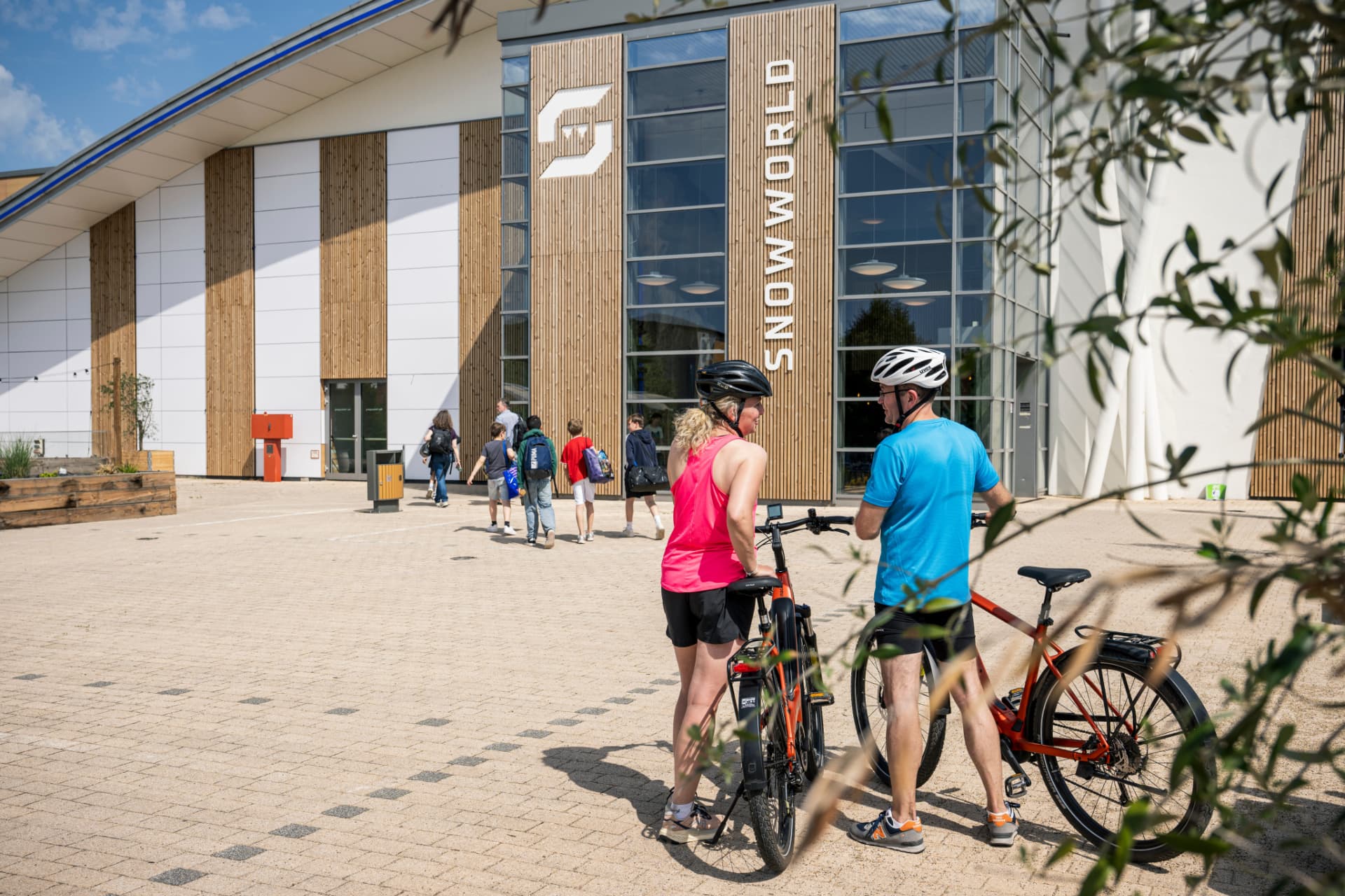 Zwei Radfahrer mit Helmen stehen im Sommer vor dem Gebäude der SnowWorld.