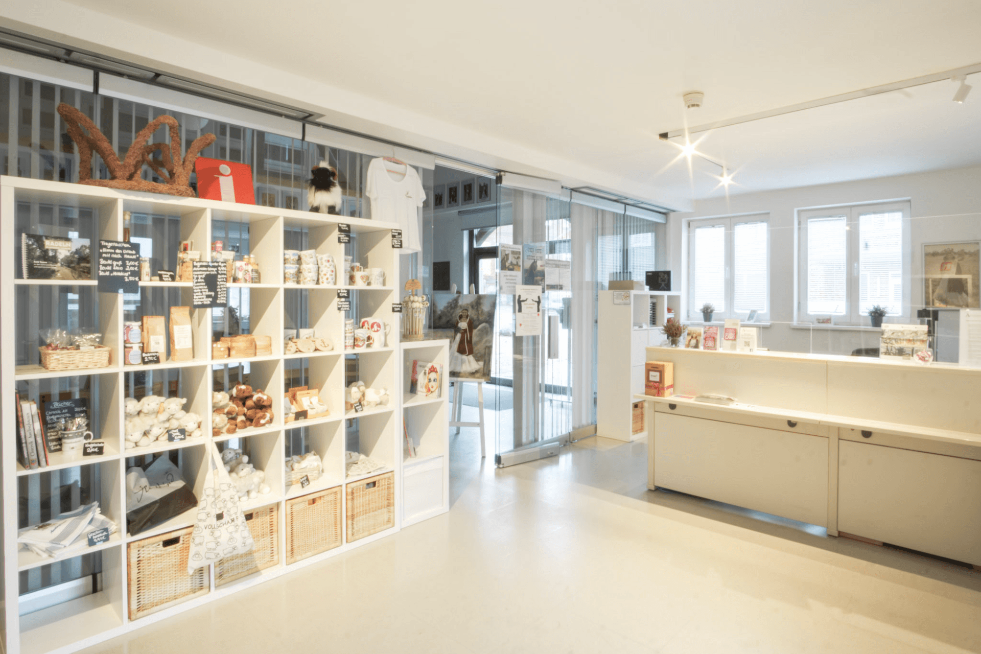 Ein moderner Empfangsbereich mit weißen Regalen voller Souvenirs, einem Tresen und großen Fenstern.